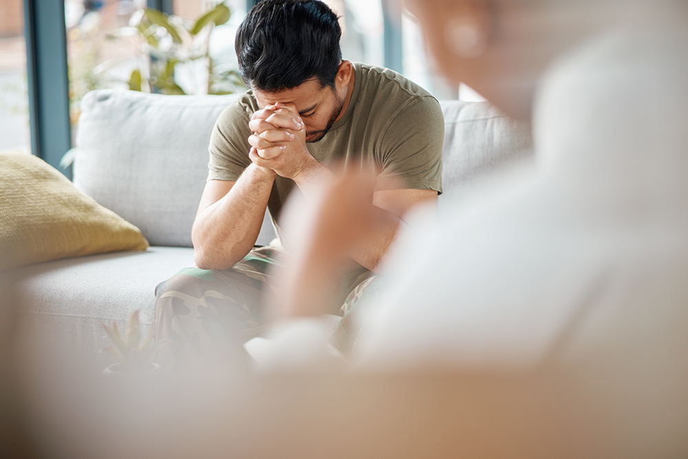 A man sitting on a couch with his hands clasped, leaning forward, while another person sits nearby out of focus.