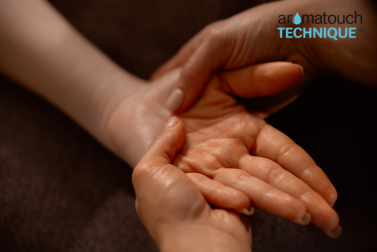 Close-up of hands gently holding another person’s hand during a touch-based wellness session, with the AromaTouch Technique logo visible.